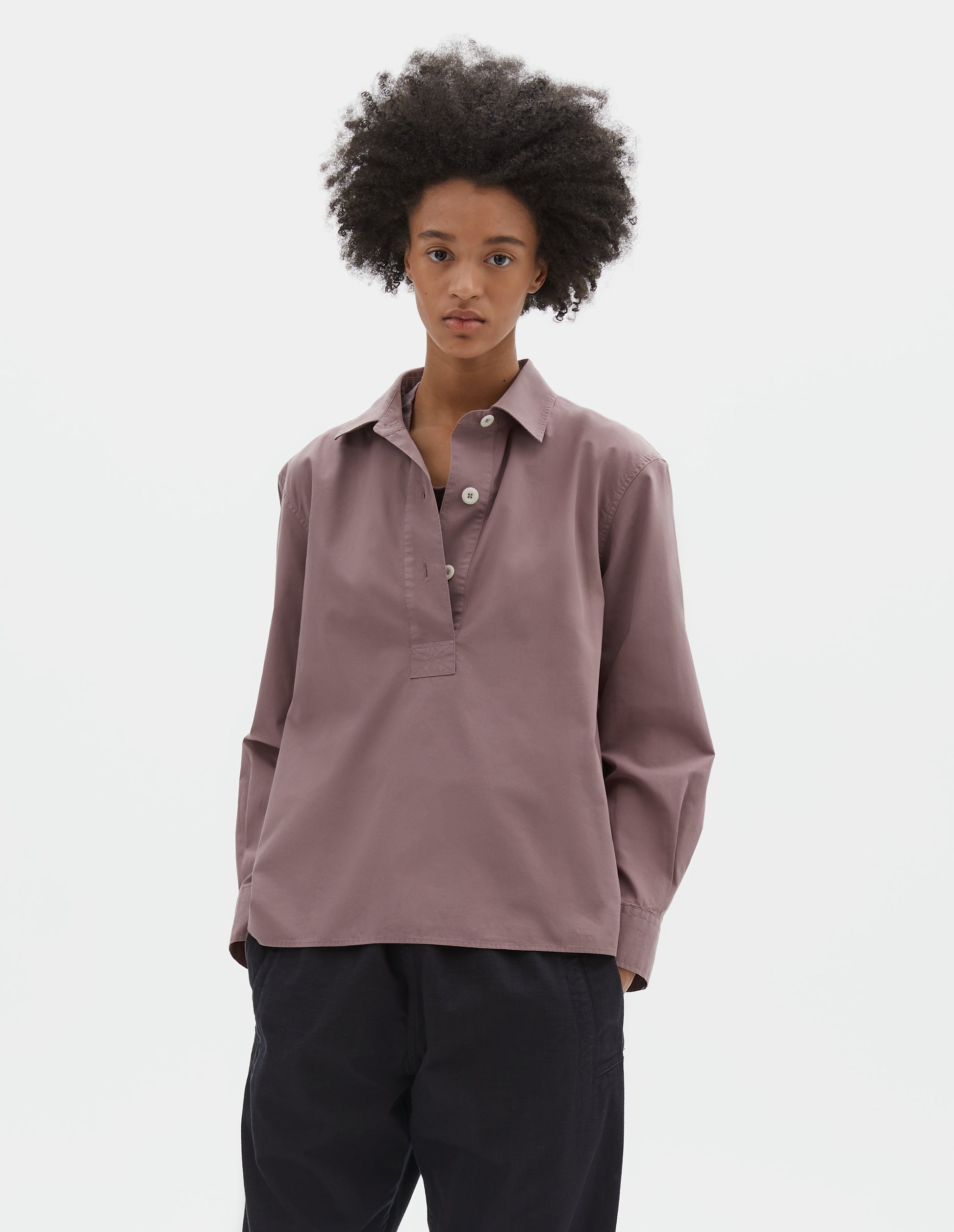 Cropped body image, front view of model wearing purpleish pink cotton poplin swing shirt, with half raised placket and corozo button fastening. Styled with dark navy trousers.