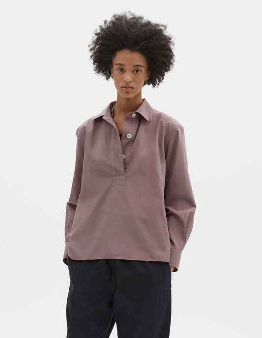 Cropped body image, front view of model wearing purpleish pink cotton poplin swing shirt, with half raised placket and corozo button fastening. Styled with dark navy trousers.