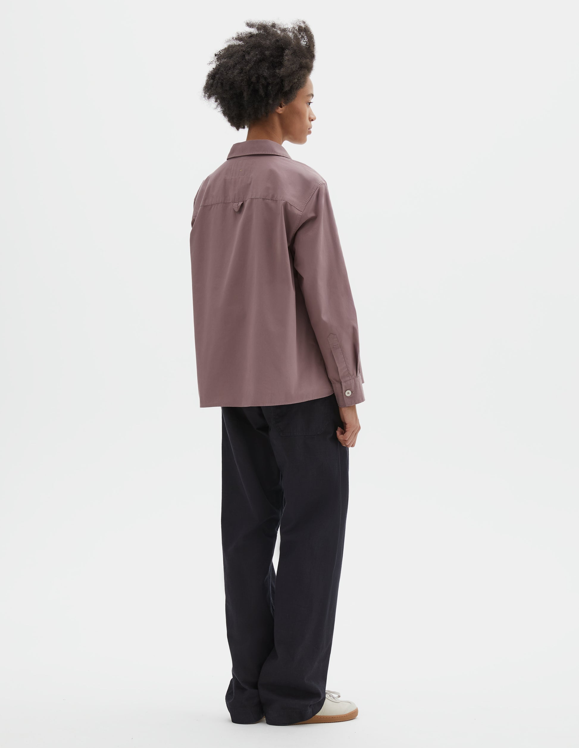 Full body image, back view of model wearing purpleish pink cotton poplin swing shirt, with corozo button cuffs and contrast hanger loop detailing on back yoke. Styled with dark navy trousers and beige suede trainers.