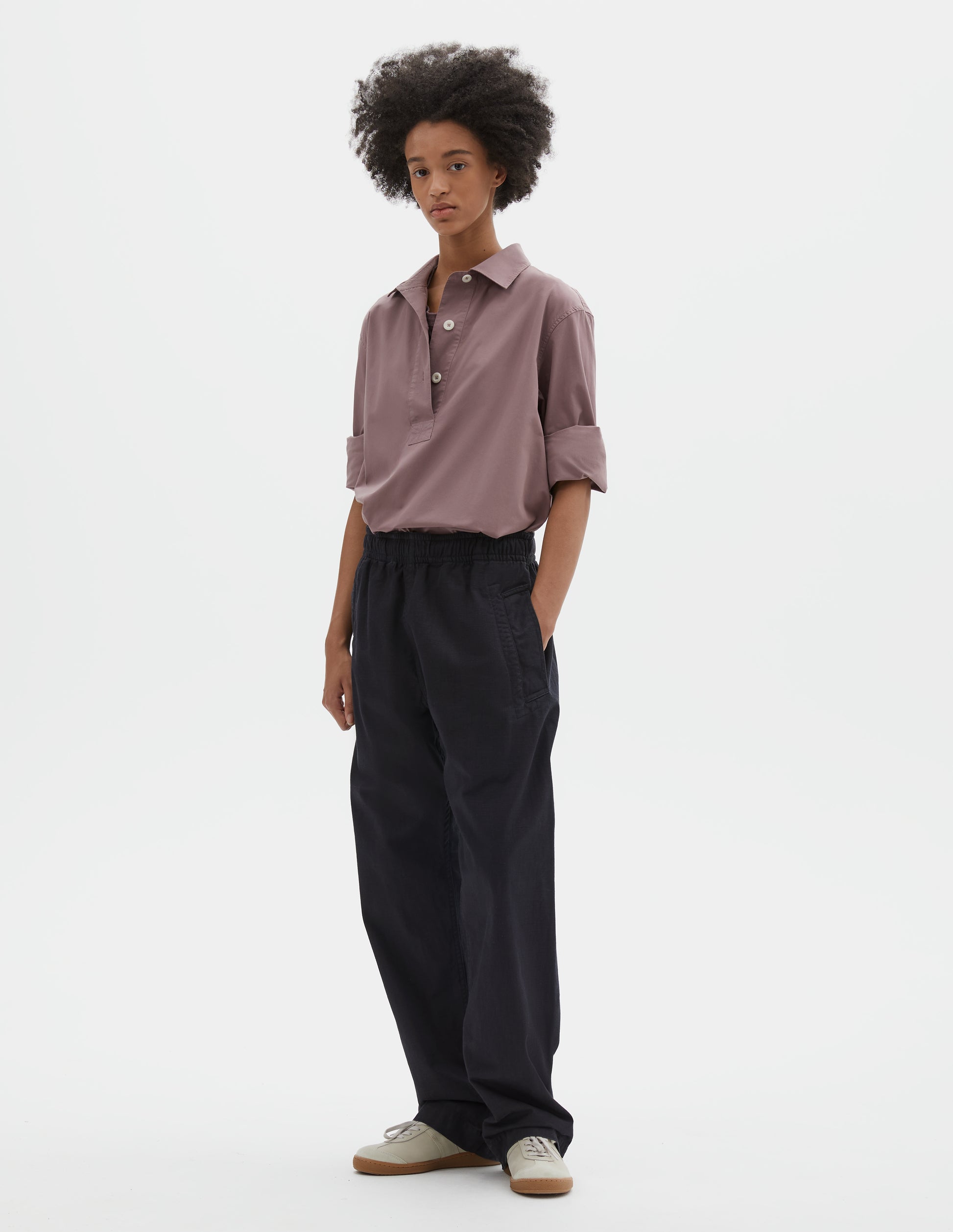 Full body image, front view of model wearing relaxed dark blue joggers with elasticated waist and deep welt pockets. Styled with lilac shirt tucked in and beige suede trainers.