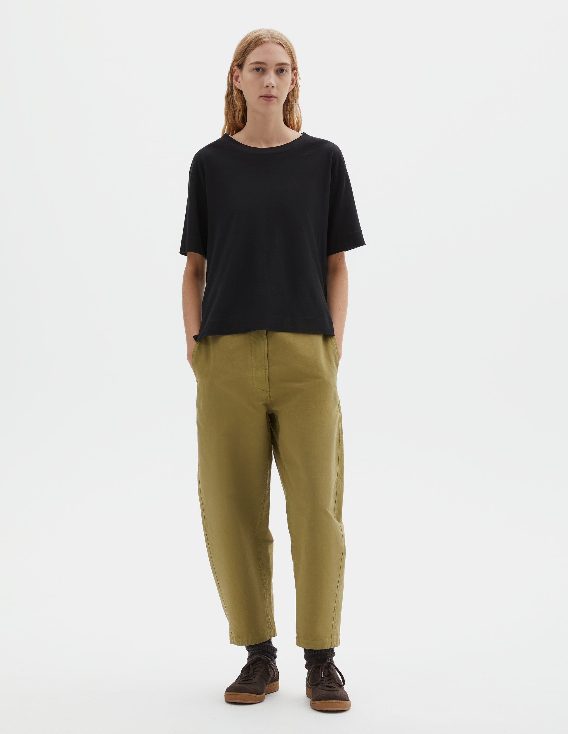 Full body image, front view of model wearing black short sleeve cotton t-shirt with contrast rib on neck. Styled with green trousers, black socks and brown suede trainers.