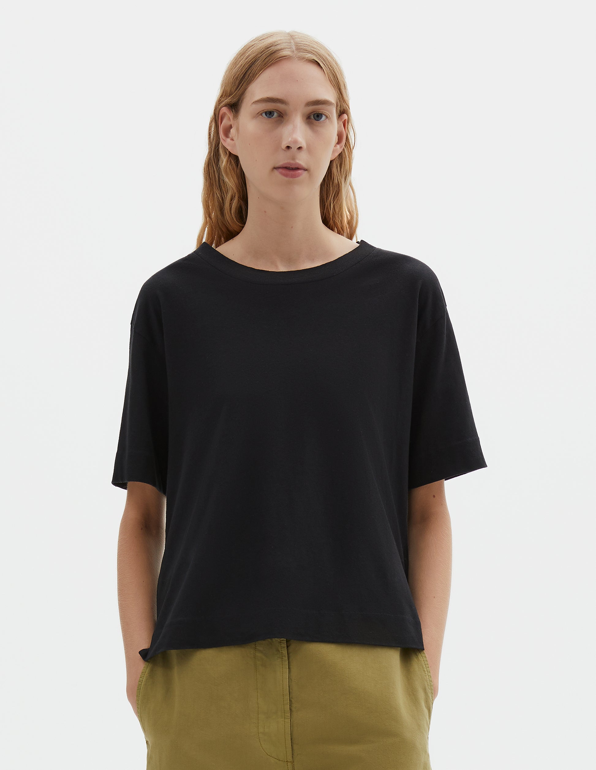 Cropped body image, front view of model wearing black short sleeve cotton t-shirt with contrast rib on neck. Styled with green trousers.