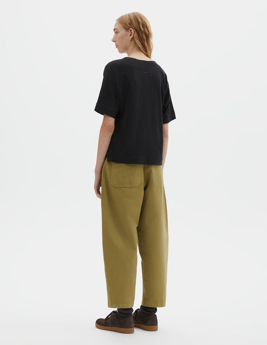 Full body image, back view of model wearing black short sleeve cotton t-shirt with contrast rib on neck and stitch detailing on back shoulder. Styled with green trousers, black socks and brown suede trainers.