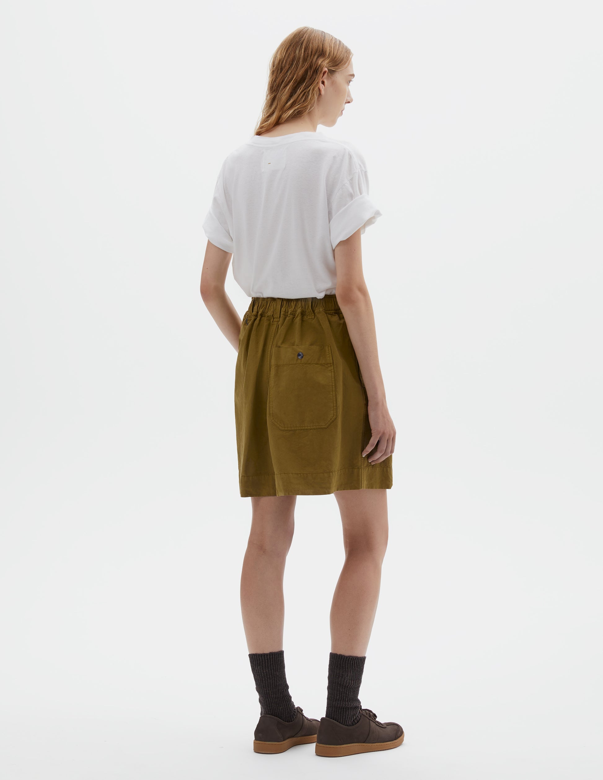 Full body image, back view of model wearing yellowish green cotton linen shorts with semi-elasticated waistband, press stud fastening on back pocket and belt loops with one D-ring detail. Styled with white t-shirt, dark brown socks and dark brown nubuck trainers.