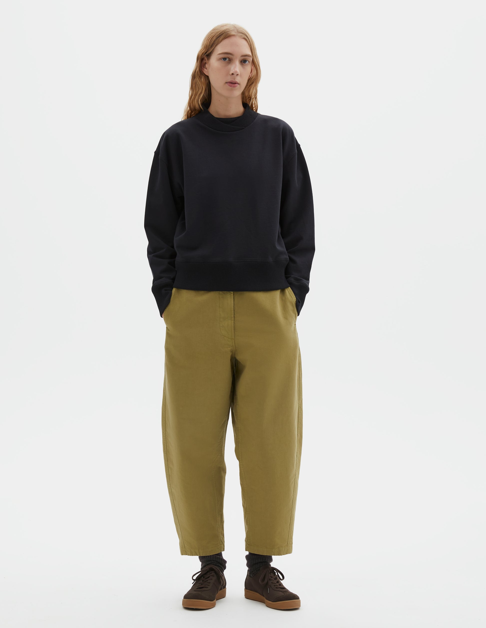 Full body image, front view of model wearing black cotton sweatshirt, with soft rib neck with crossover detail and ribbed cuffs and hem. Styled with light green trousers, brown socks and brown suede trainers.