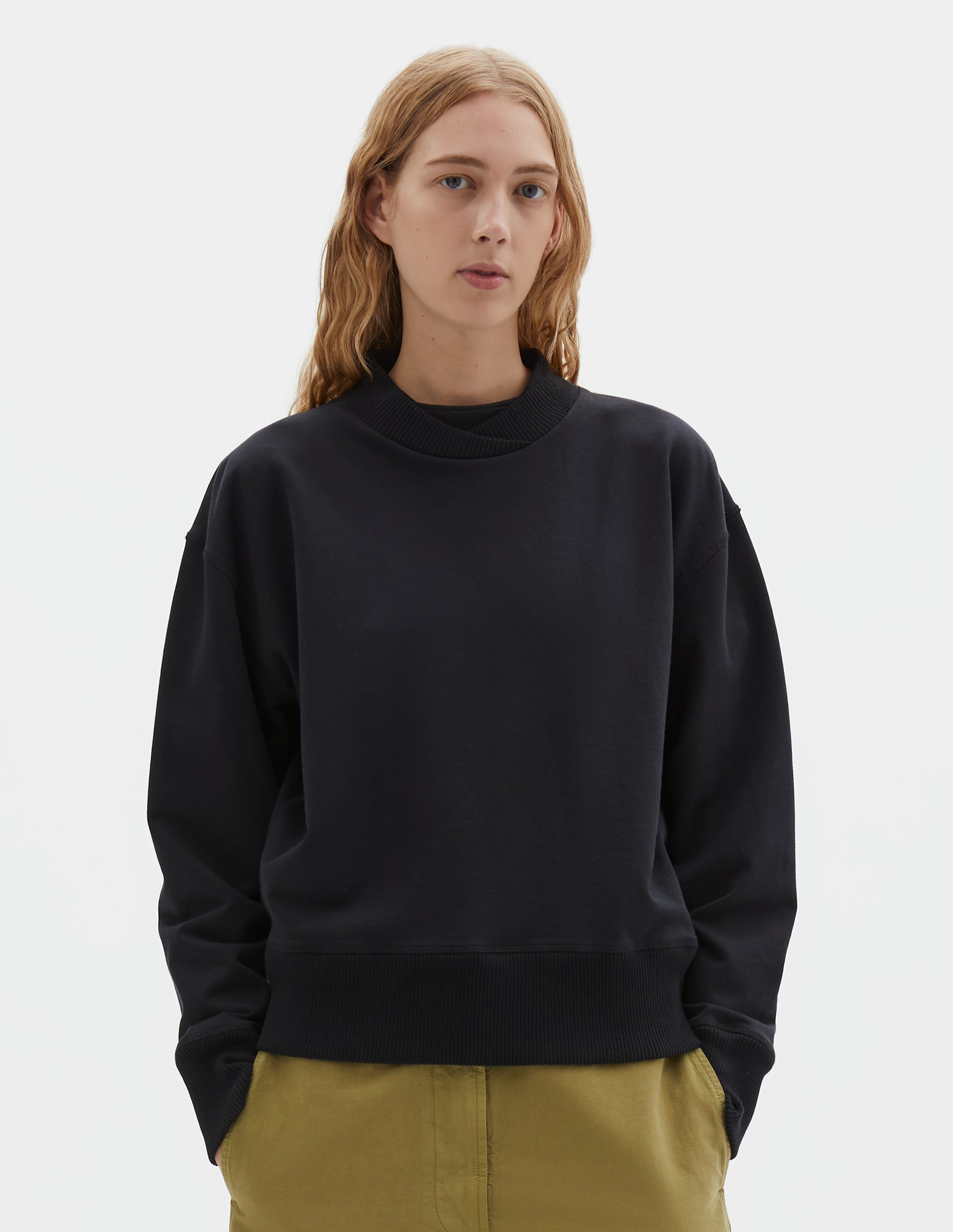Cropped body image, front view of model wearing black cotton sweatshirt, with soft rib neck with crossover detail and ribbed cuffs and hem. Styled with light green trousers.