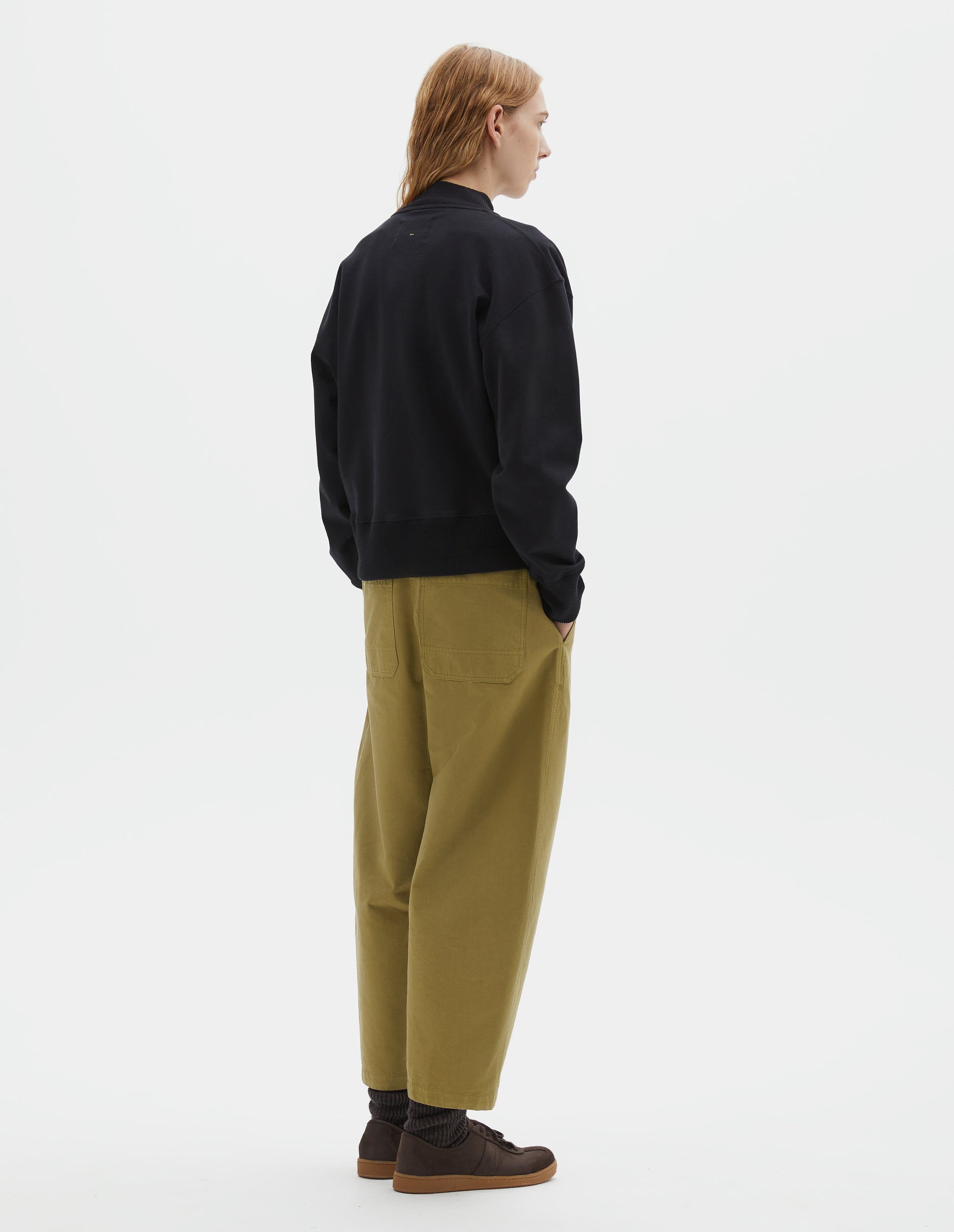 Full body image, back view of model wearing black cotton sweatshirt, with ribbed neck, cuffs and hem. Styled with light green trousers, brown socks and brown suede trainers.