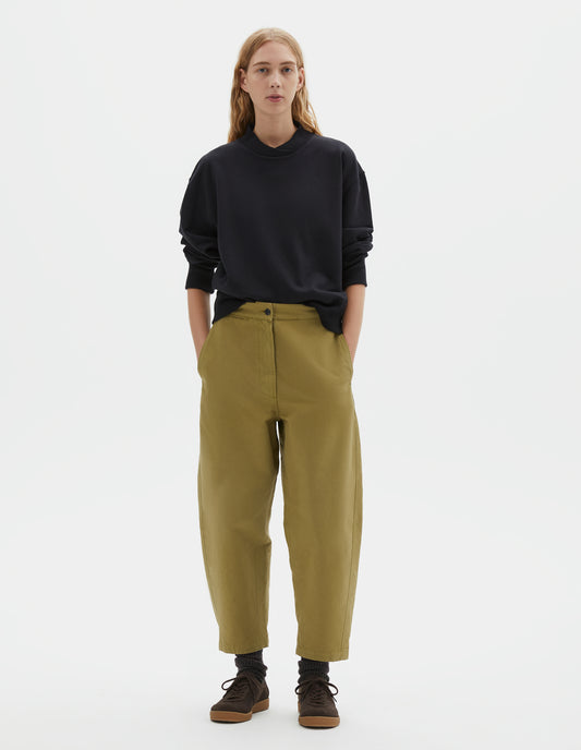 Full body image, front view of model wearing cropped light green barrel leg trousers with slant side pockets, silver metal closure and adjustable waist tab detailing. Styled with black jumper, dark brown socks and dark brown nubuck trainers.