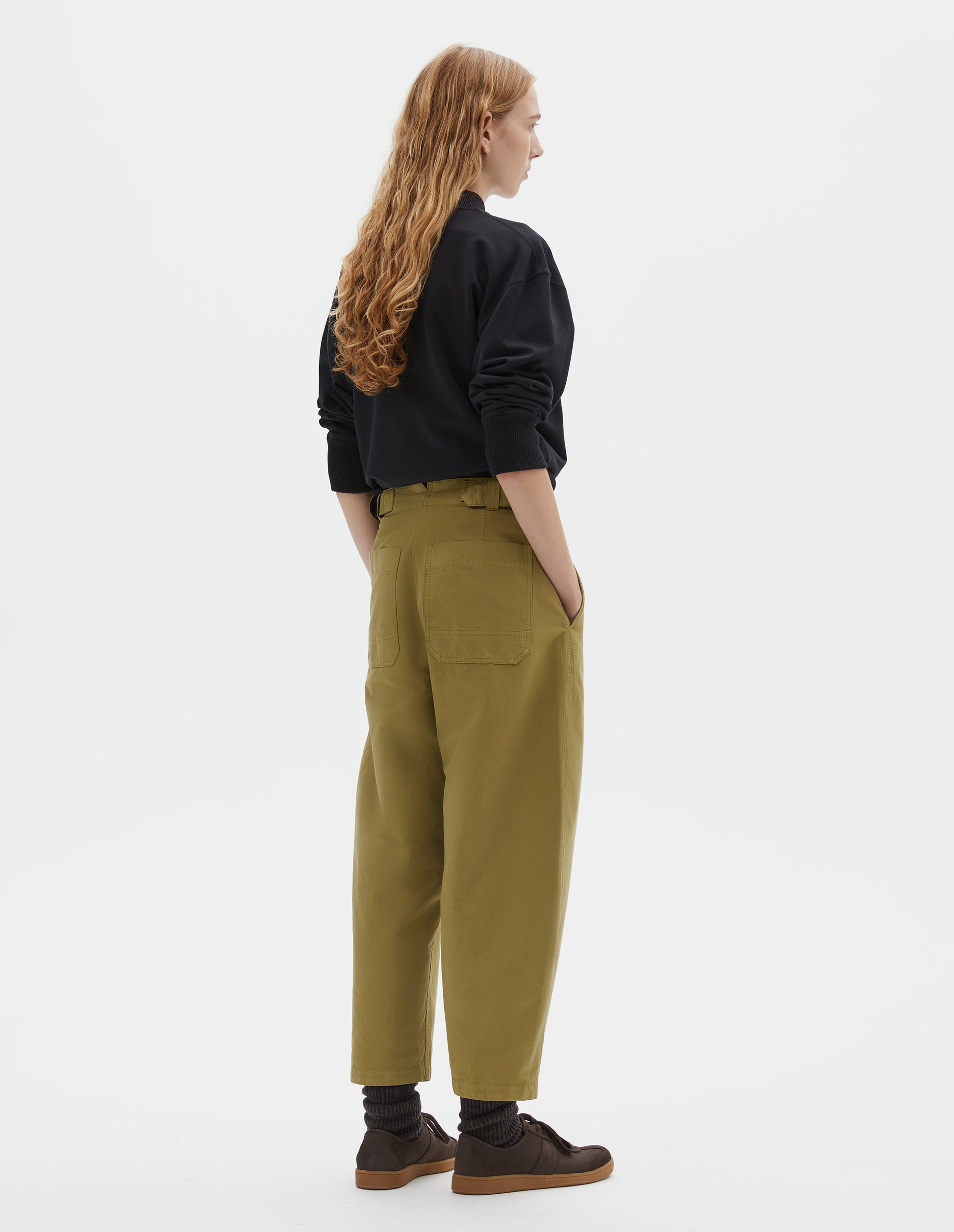 Full body image, back view of model wearing cropped light green barrel leg trousers with two patch pockets and silver metal waist tab adjuster. Styled with black jumper, dark brown socks and dark brown nubuck trainers.