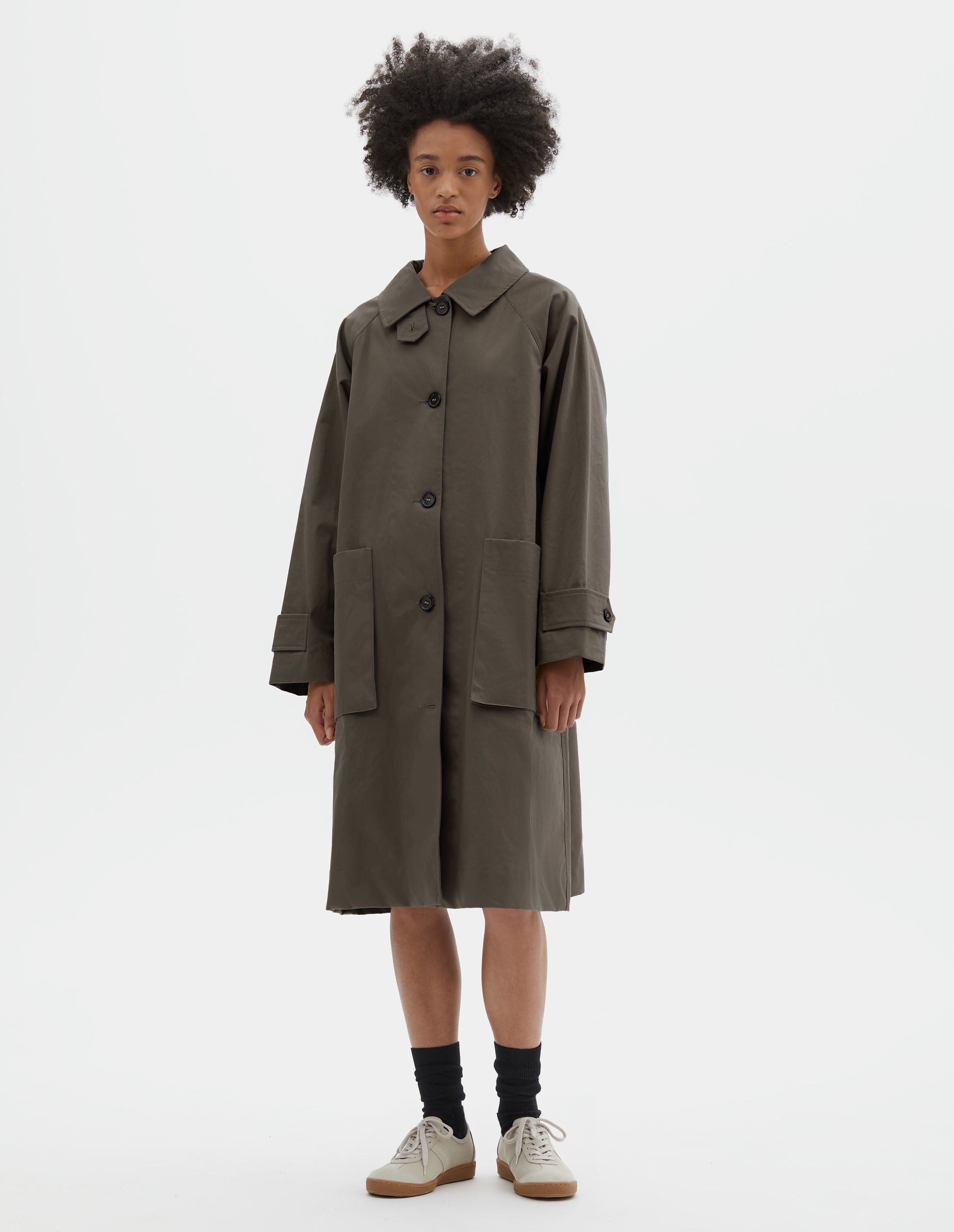 Full body image, front view of model wearing greyish brown cotton canvas long sleeve mac, with storm tab fastening neck, raglan sleeve, button tab adjustable cuffs and large patch bellow pockets. Styled over white t-shirt and navy blue shorts, with black socks and off white suede trainers.