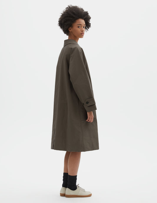 Full body image, back view of model wearing greyish brown cotton canvas long sleeve mac, with button tab adjustable cuffs and rear vent. Styled with black socks and off white suede trainers.