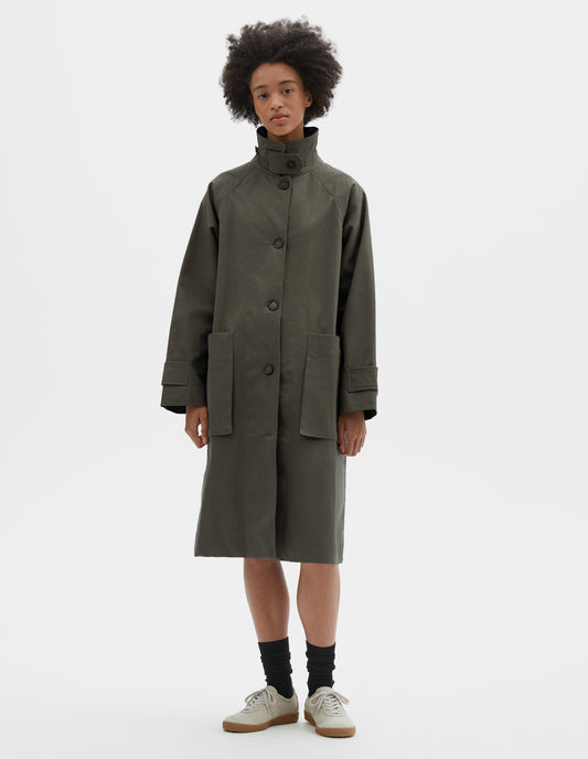 Full body image, front view of model wearing greyish brown cotton canvas long sleeve mac, with storm tab fastening neck stood up, raglan sleeve, button tab adjustable cuffs and large patch bellow pockets. Styled with the collar popped, over white t-shirt and navy blue shorts, with black socks and off white suede trainers.