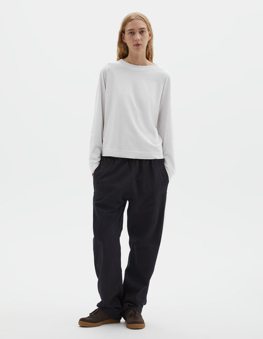 Full body image, front view of model wearing white cotton linen long sleeve t-shirt with contrast rib on neckline and raglan sleeve detailing. Styled with dark navy joggers and brown suede trainers.