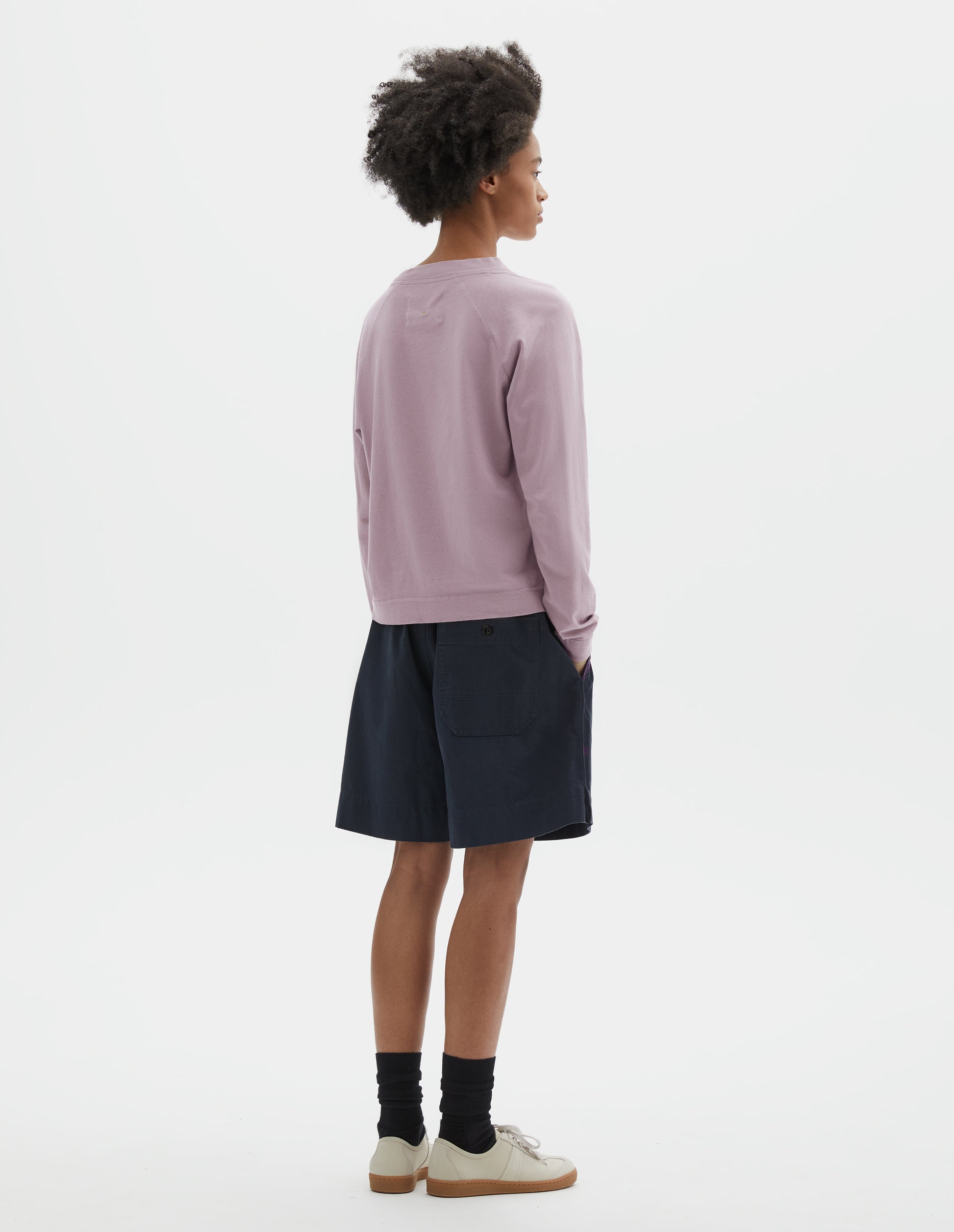 Fully body image, back view of model wearing dark blue cotton shorts with elasticated waist, concealed side pockets, herringbone tape drawcord and side split hem. Styled with lilac long sleeved t shirt, black socks and white suede shoes with gum sole.