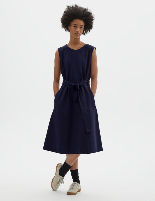 Full body image, front view of model wearing dark navy cotton sleeveless dress with round neck, wrap around belt, side seam pockets and deep stitch hem. Styled with black socks and beige suede trainers.