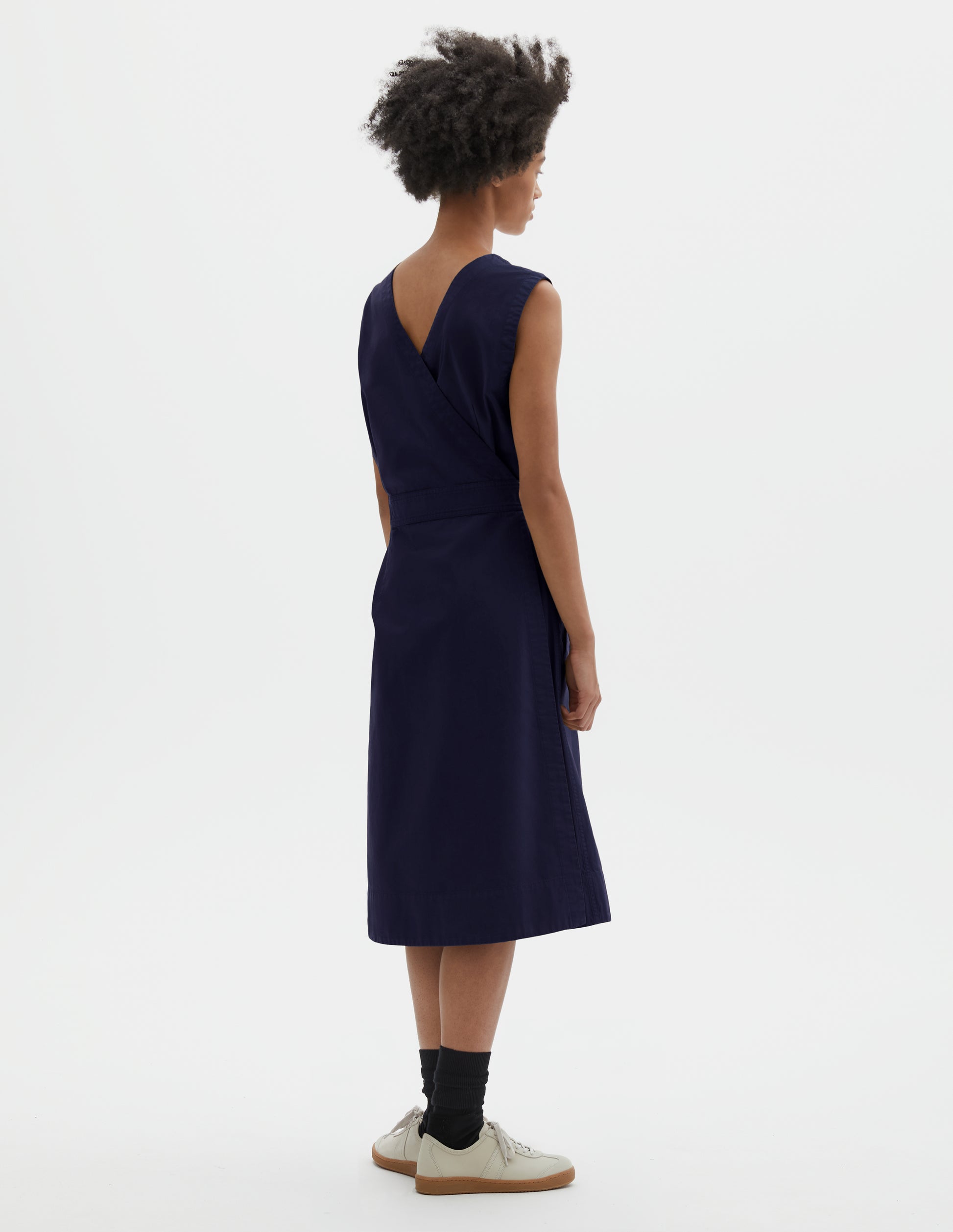 Full body image, back view of model wearing dark navy cotton sleeveless dress with v detail on back, wrap around belt, back patch pocket and deep stitch hem. Styled with black socks and beige suede trainers.