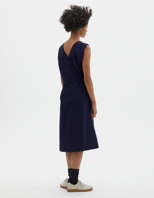 Full body image, back view of model wearing dark navy cotton sleeveless dress with v detail on back, wrap around belt, back patch pocket and deep stitch hem. Styled with black socks and beige suede trainers.