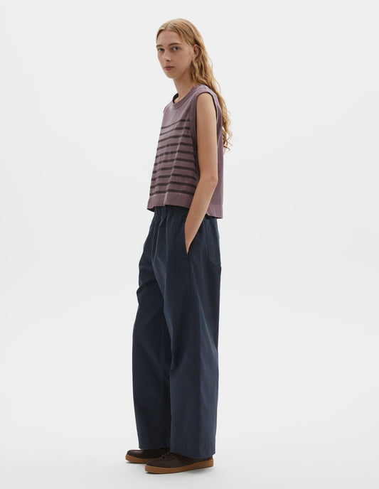 Full body image, front view of model wearing purple cotton linen tank top with brown stripes, contrast ribbing on neckline and armholes, with side split hem and deep stitch hem. Styled with dark blue trousers and brown suede trainers with gum sole. 