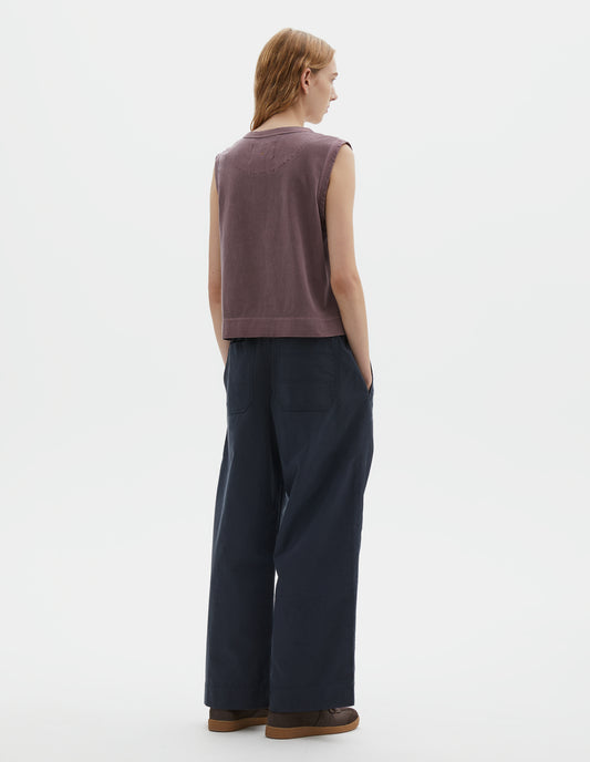 Full body image back view of model wearing purple cotton linen tank top with contrast ribbing on neckline and armholes, with stitch-through half moon on back yoke, side splits and deep stitch hem. Styled with dark blue trousers and brown suede trainers with gum sole. 