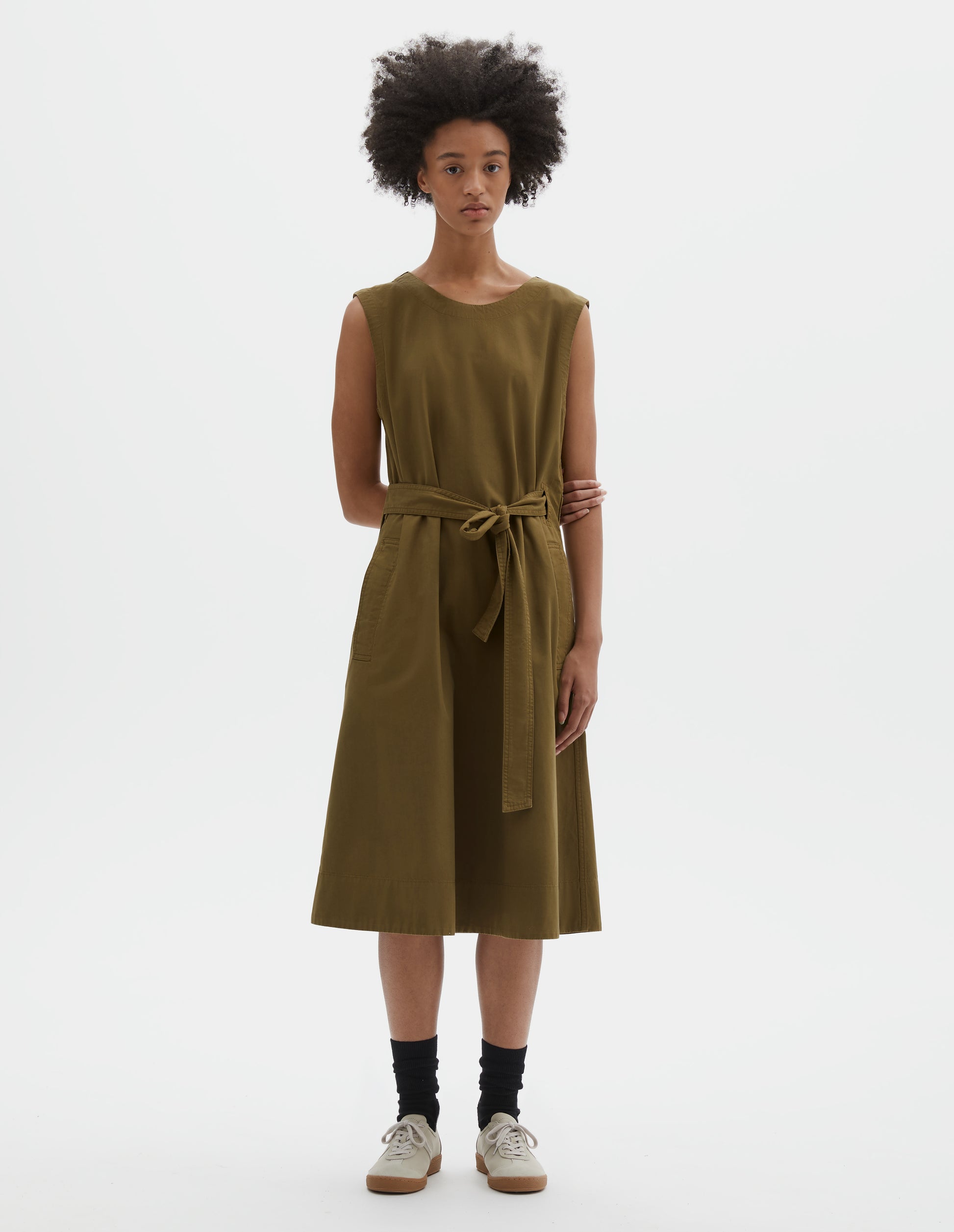 Full body image, front view of model wearing green cotton sleeveless dress with round neck, wrap around belt, side seam pockets and deep stitch hem. Styled with black socks and beige suede trainers.