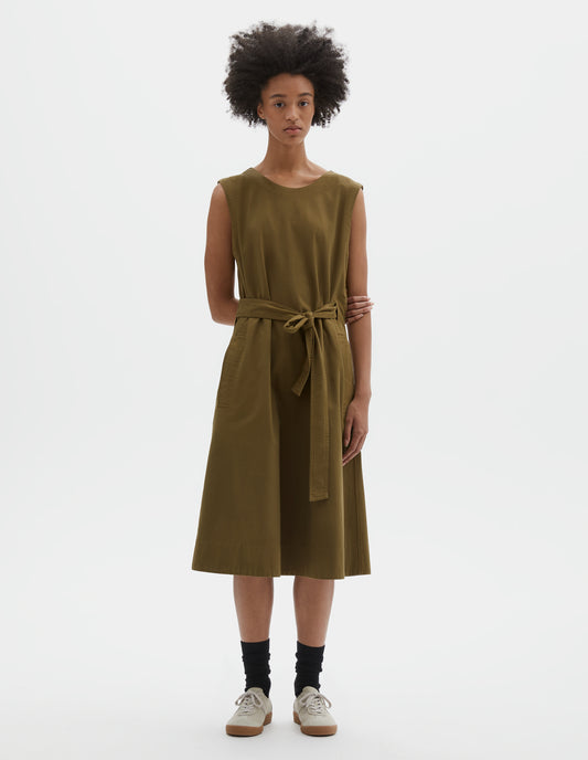 Full body image, front view of model wearing green cotton sleeveless dress with round neck, wrap around belt, side seam pockets and deep stitch hem. Styled with black socks and beige suede trainers.