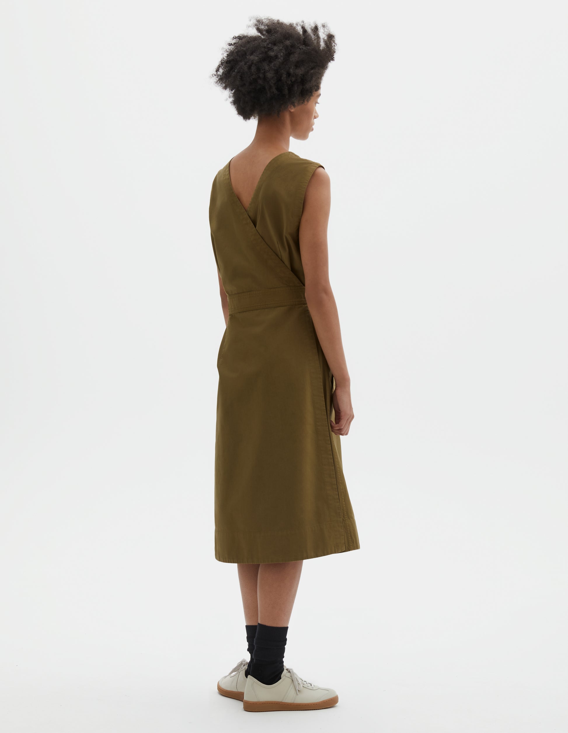 Full body image, back view of model wearing green cotton sleeveless dress with v detail on back, wrap around belt, back patch pocket and deep stitch hem. Styled with black socks and beige suede trainers.
