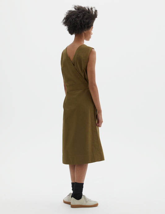 Full body image, back view of model wearing green cotton sleeveless dress with v detail on back, wrap around belt and deep stitch hem. Styled with black socks and beige suede trainers.
