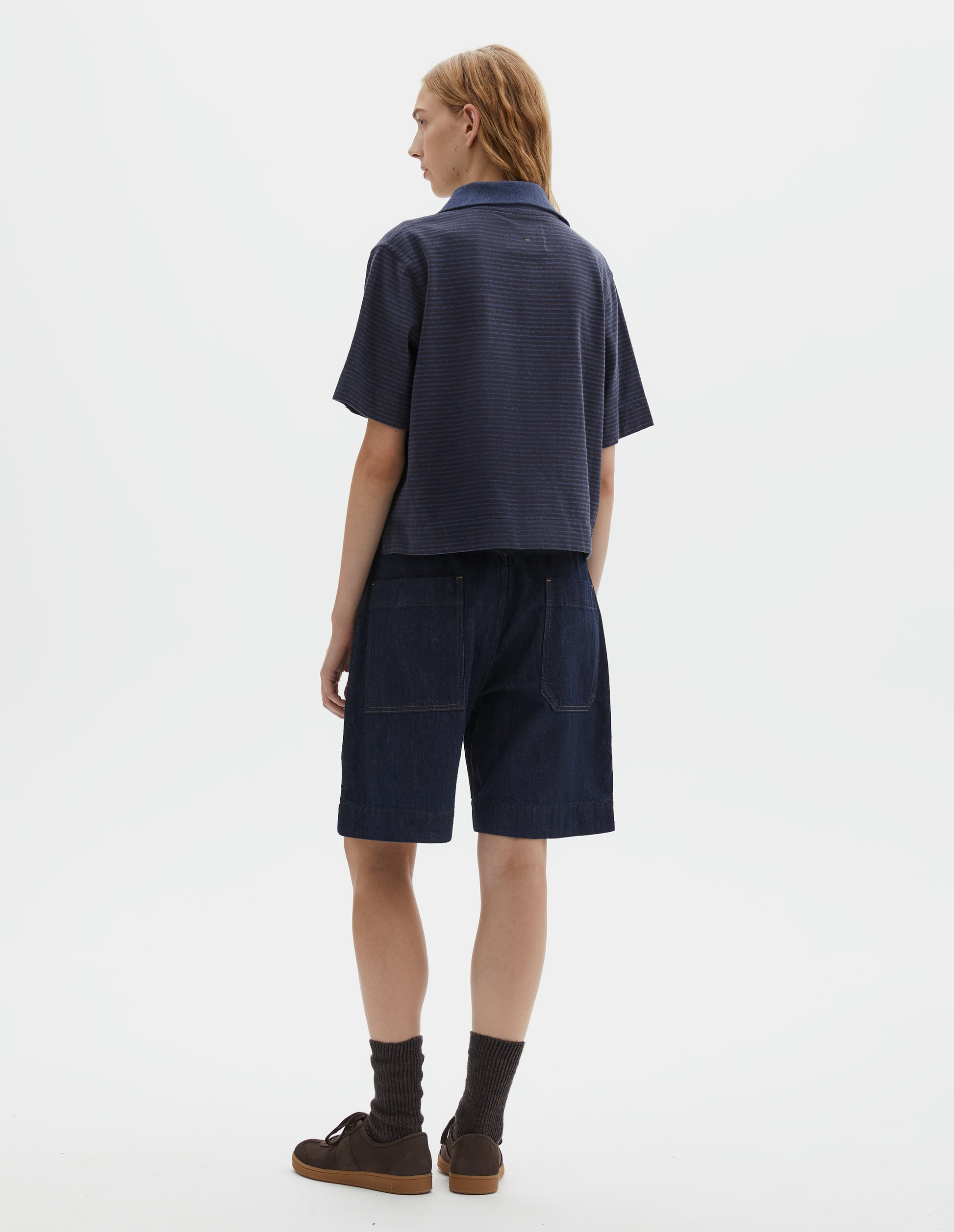 Full body image, back view of model wearing dark blue and light blue striped polo t-shirt, with contrast melange rib collar and side split hem. Styled with indigo blue denim shorts, brown socks and brown suede trainers.