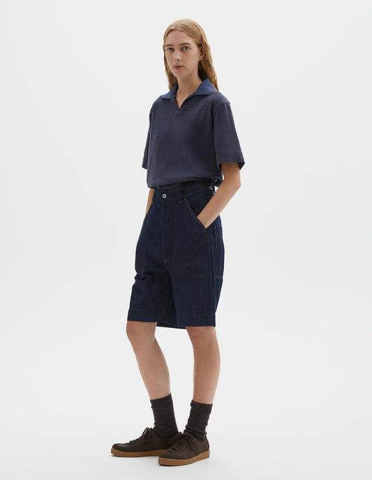 Full body image, front view of model wearing dark blue relaxed denim twill shorts with metal button tab waist adjuster, metal button fly, belt loops and slant stitch-through pockets. Styled with blue polo, dark brown socks and dark brown nubuck trainers.