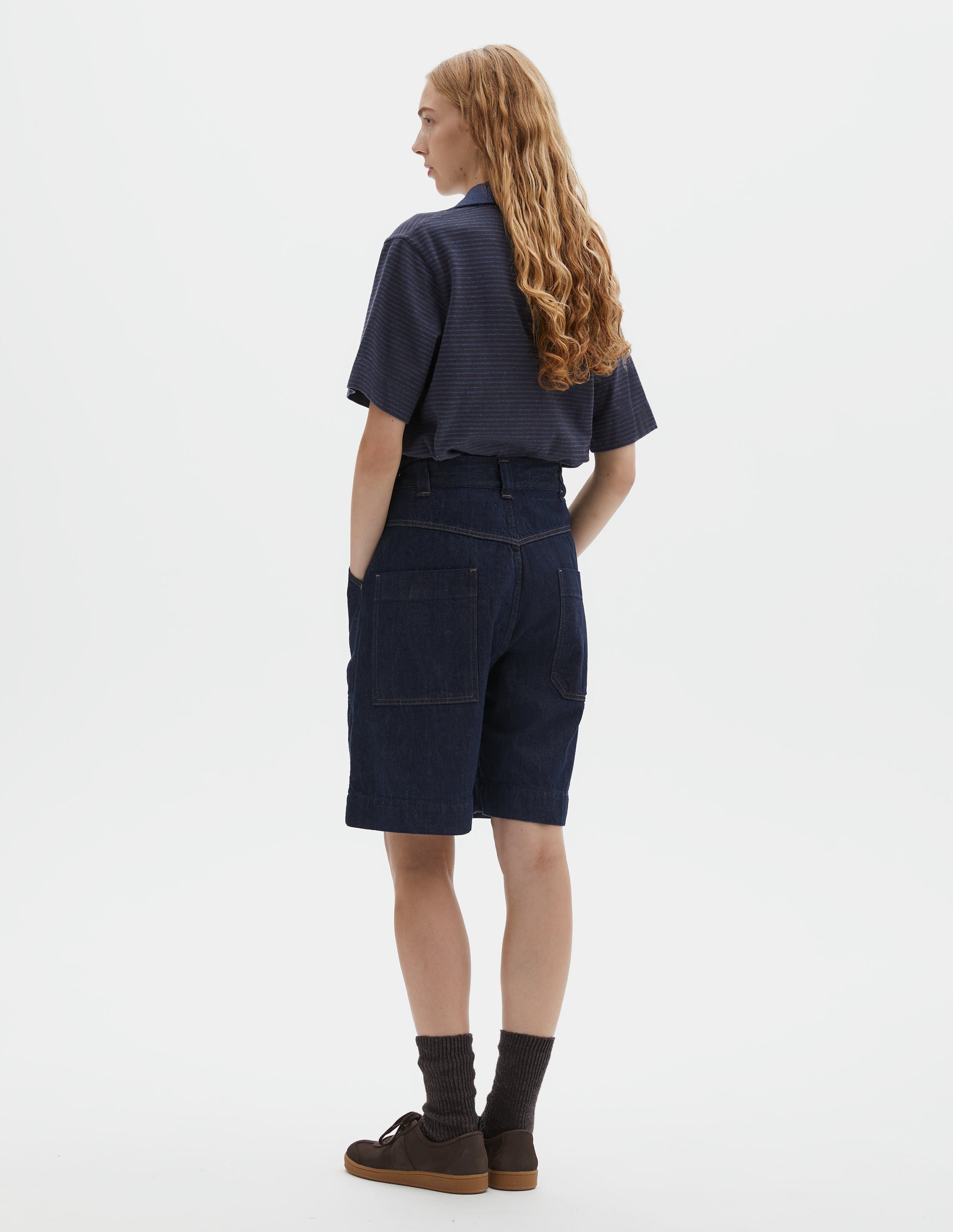 Full body image, back view of model wearing dark blue relaxed denim twill shorts with metal button tab waist adjusters, belt loops and patch pockets. Styled with blue polo, dark brown socks and dark brown nubuck trainers.