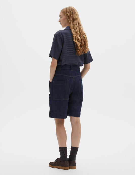 Full body image, back view of model wearing dark blue relaxed denim twill shorts with metal button tab waist adjusters, belt loops and patch pockets. Styled with blue polo, dark brown socks and dark brown nubuck trainers.