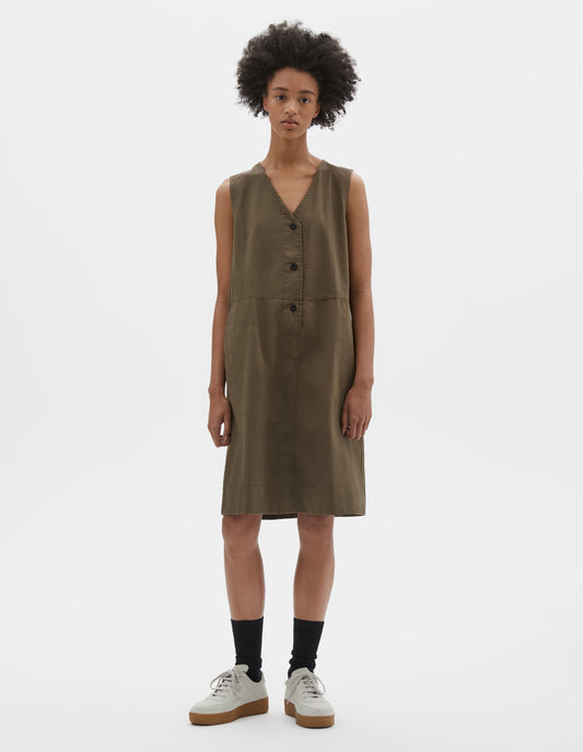 Full body image, front view of model wearing greenish brown sleeveless dress with v neck detailing, slant side pockets and corozo brace button closure. Styled with black socks and beige suede trainers.