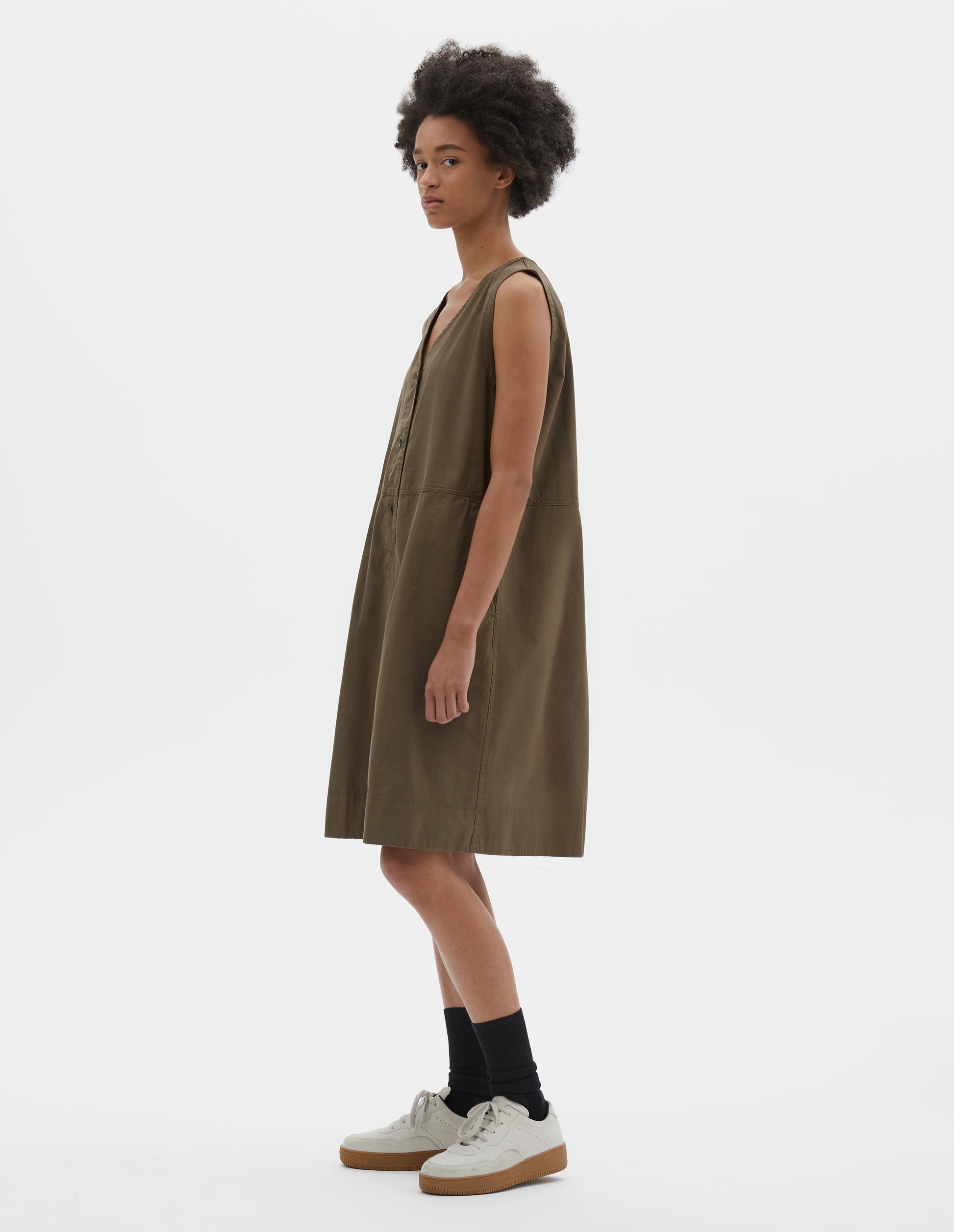 Full body image, side view of model wearing greenish brown sleeveless dress with v neck detailing, slant side pockets and corozo brace button closure. Styled with black socks and beige suede trainers.