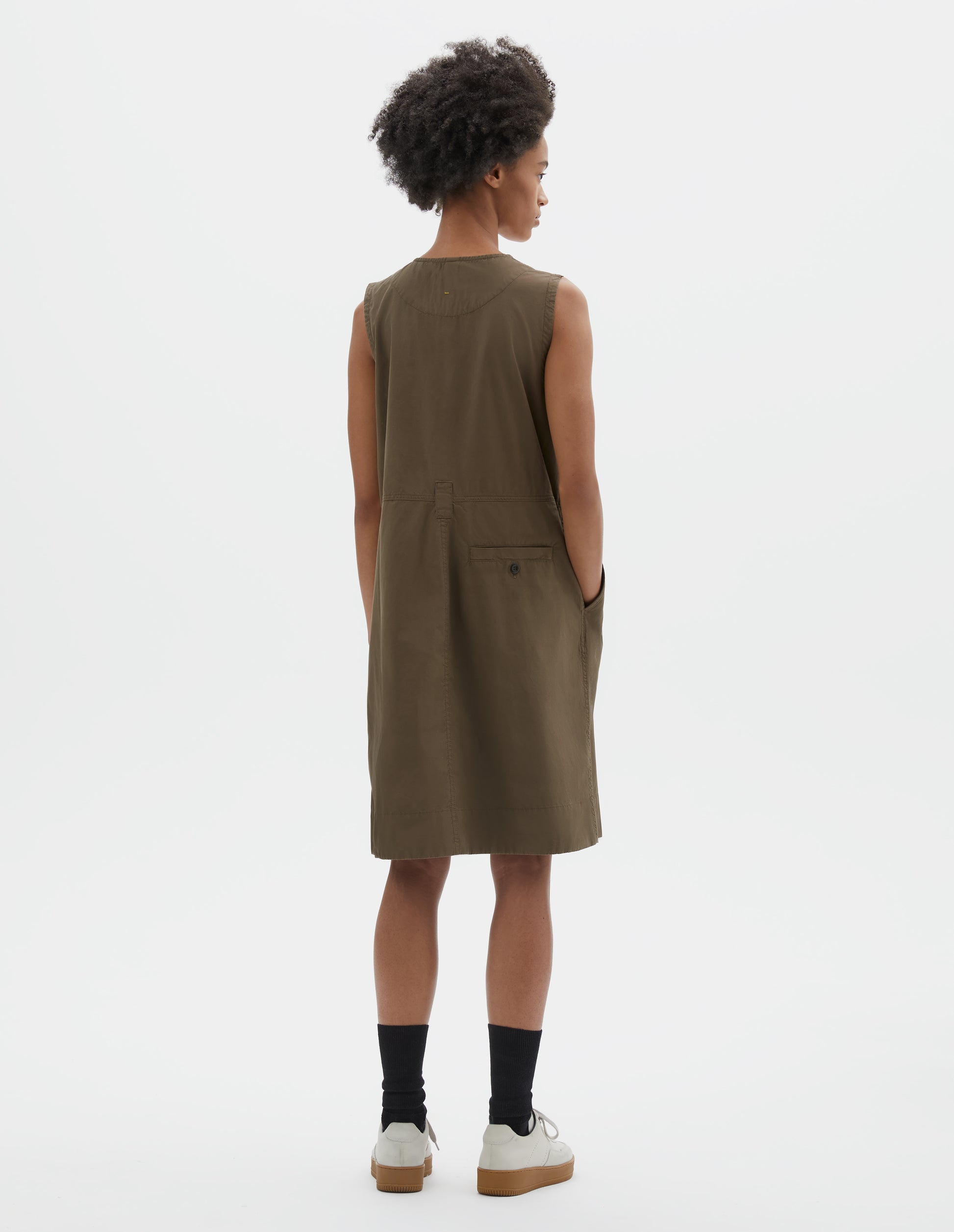 Full body image, back view of model wearing greenish brown sleeveless dress with singular belt loop and back button through welt pocket. Styled with black socks and beige suede trainers.