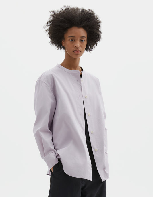 Cropped body image, front view of model wearing light grey cotton canvas collarless shirt with corozo button fastening and curved hem detailing. Styled over black t-shirt and with black trousers.