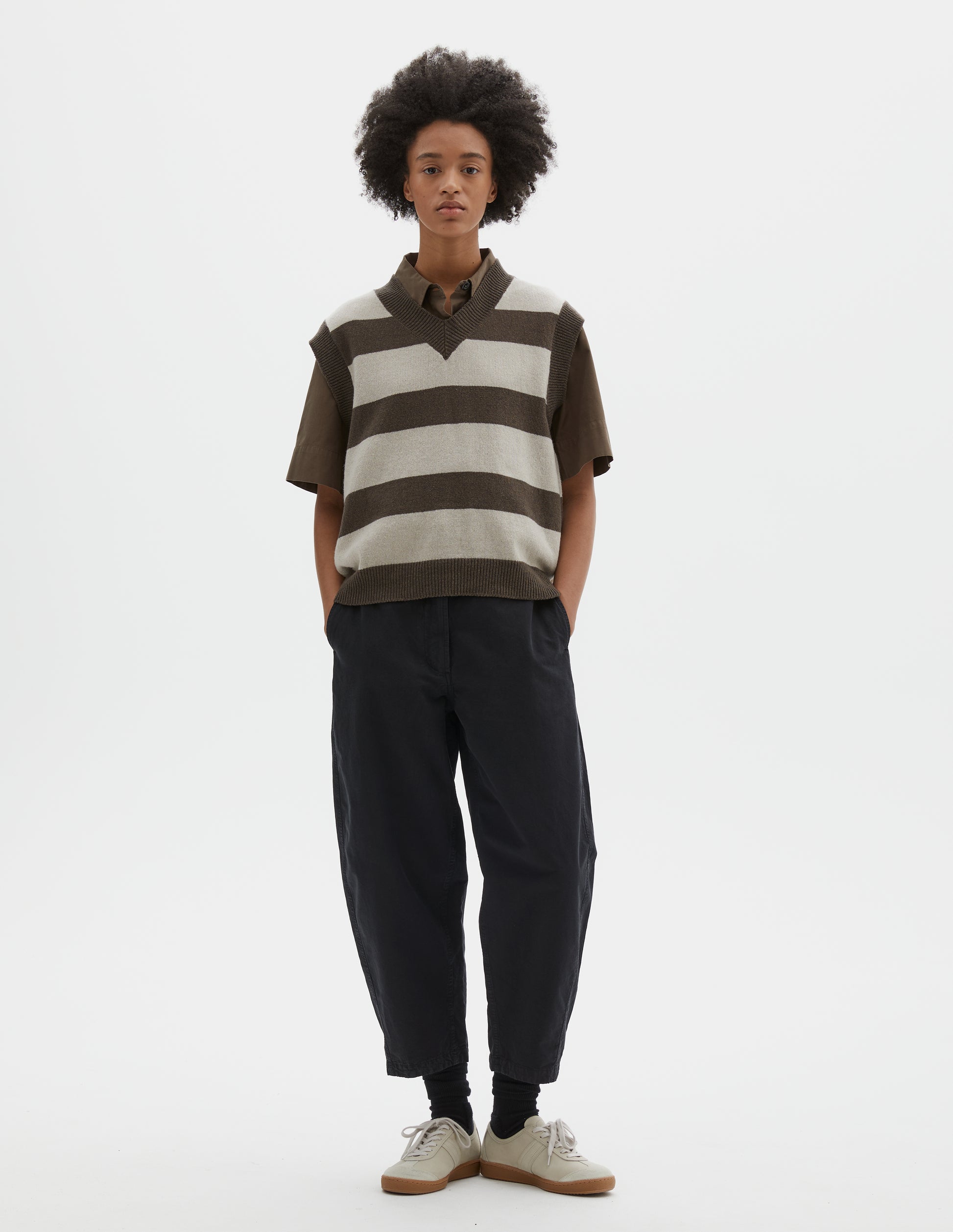 Full body image, front view of model wearing brown and off white chunky striped sleeveless knitted jumper, with thick brown rib on v neck, armholes and hem. Styled with short sleeve brown shirt and black trousers, with black socks and beige nubuck trainers.