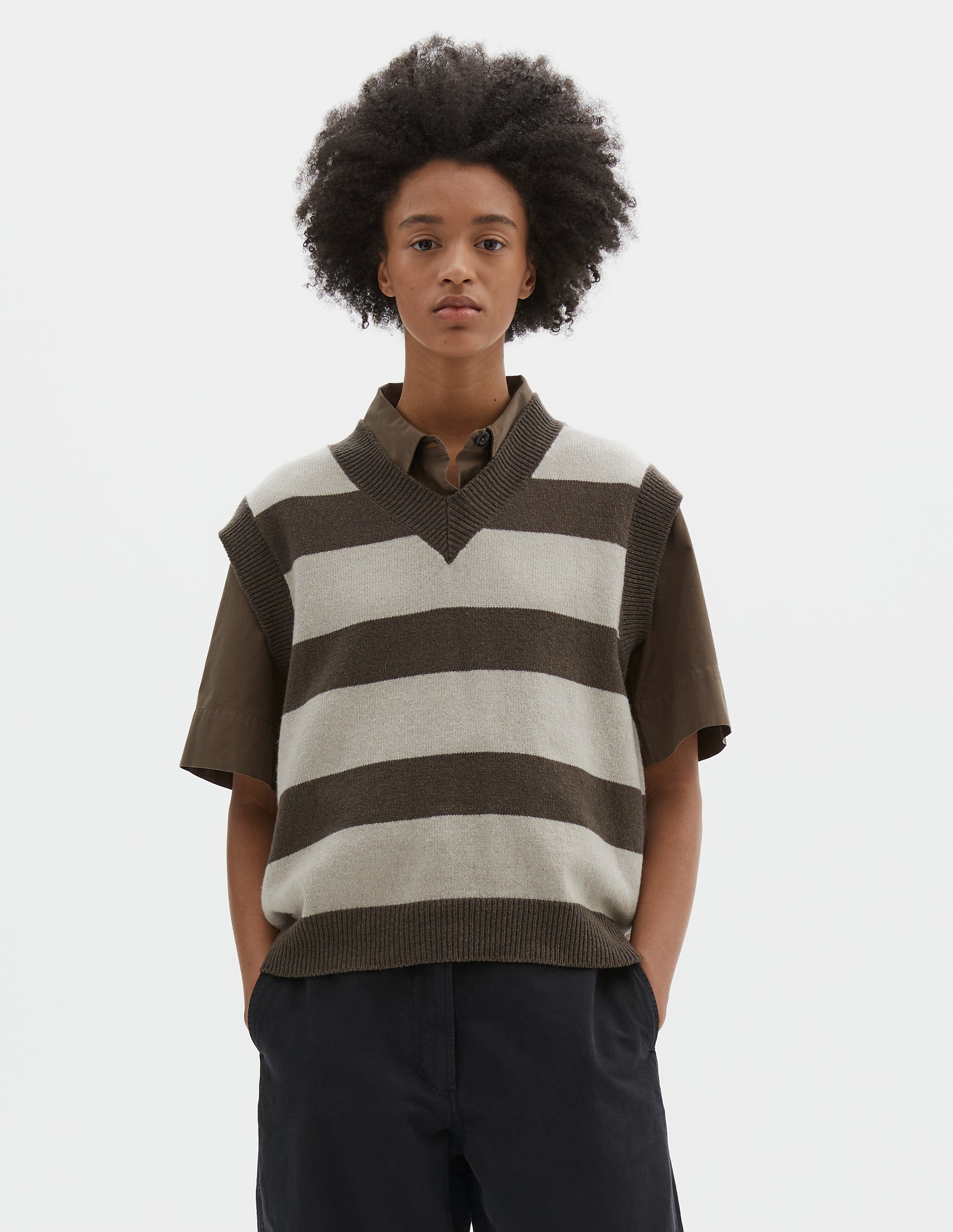 Cropped body image, front view of model wearing brown and off white chunky striped sleeveless knitted jumper, with thick brown rib on v neck, armholes and hem. Styled with short sleeve brown shirt and black trousers.