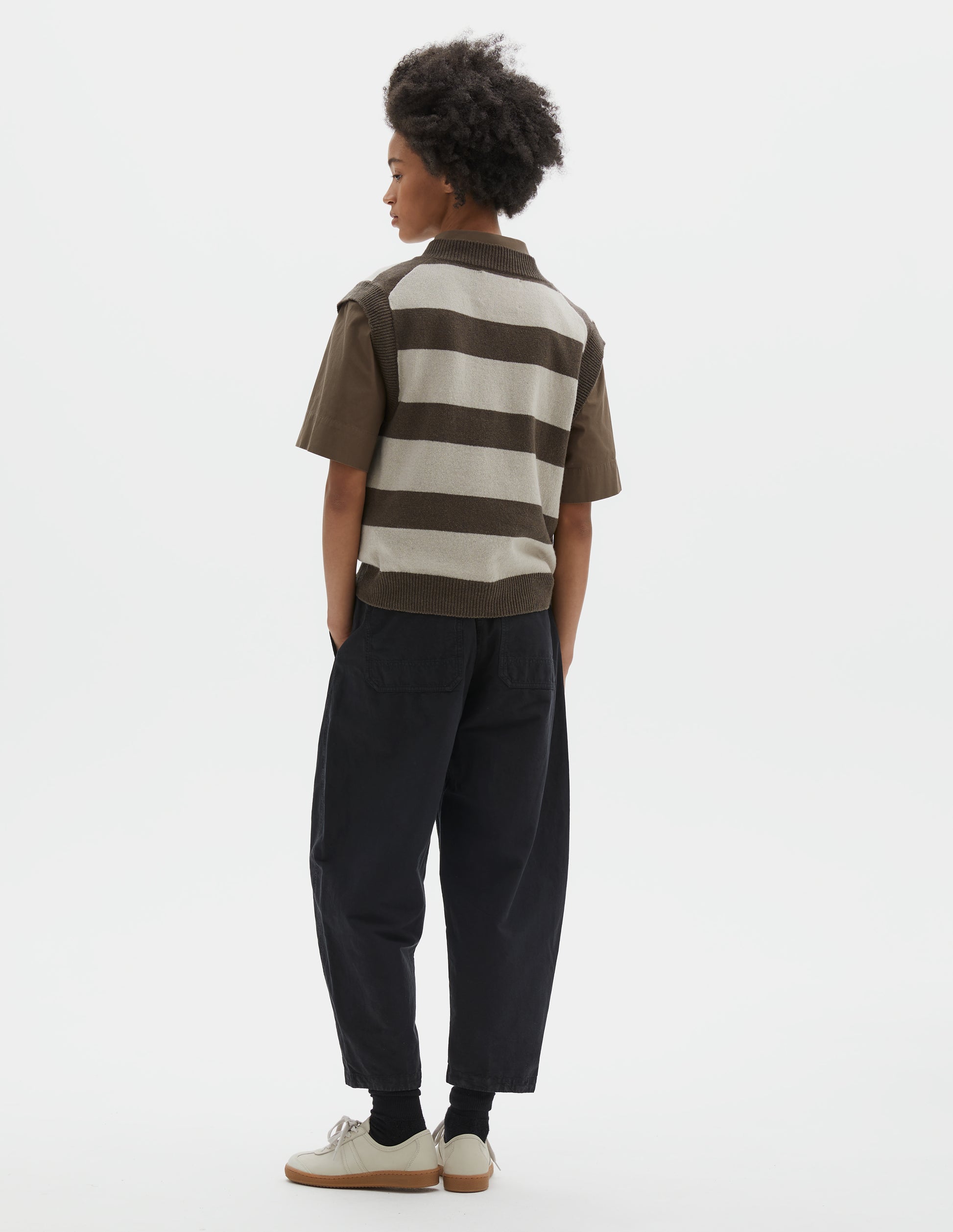 Full body image, back view of model wearing cropped black barrel leg trousers with two patch pockets and silver metal waist tab adjuster. Styled with brown short-sleeved shirt, white and brown slipover, black socks and beige nubuck trainers.