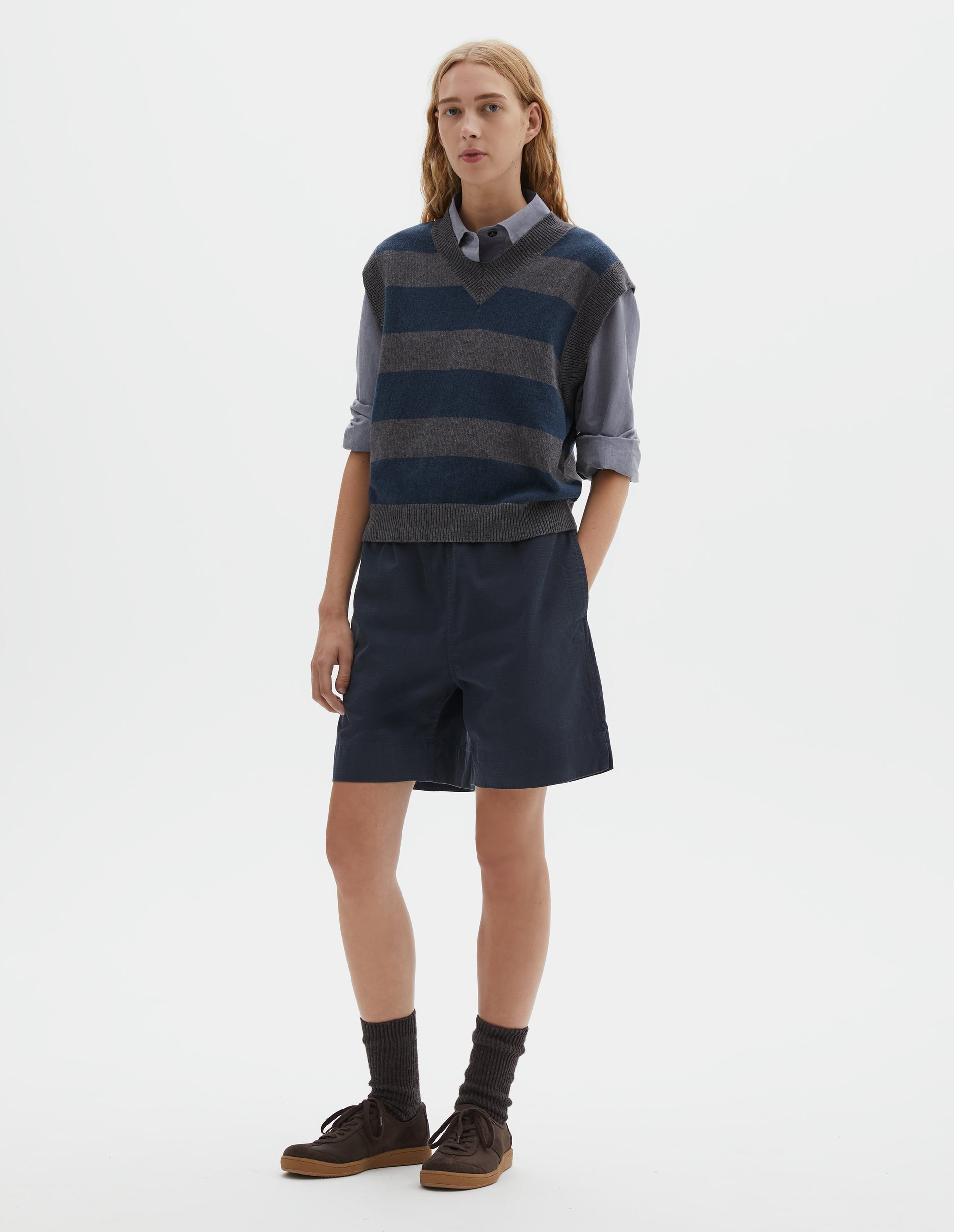 Full body image, front view of model wearing grey and dark navy striped v neck slipover, with contrast rib v neck, armholes and hem. Styled with light blue collared shirt, dark navy blue shorts, black socks and dark brown sude trainers with gum sole.