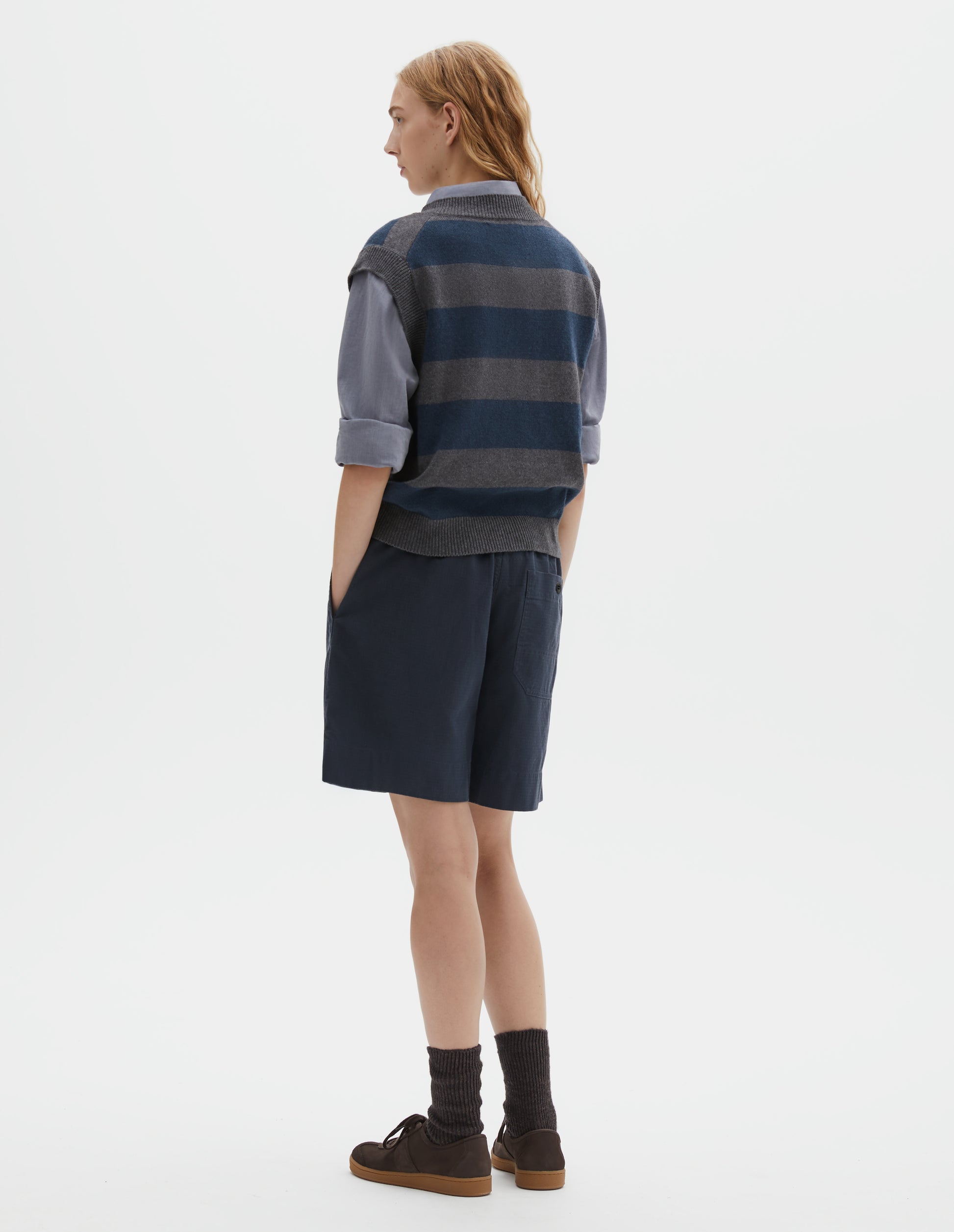 Full body image, back view of model wearing grey and dark navy striped v neck slipover, with contrast rib v neck, armholes and hem. Styled with light blue collared shirt, dark navy blue shorts, black socks and dark brown sude trainers with gum sole.