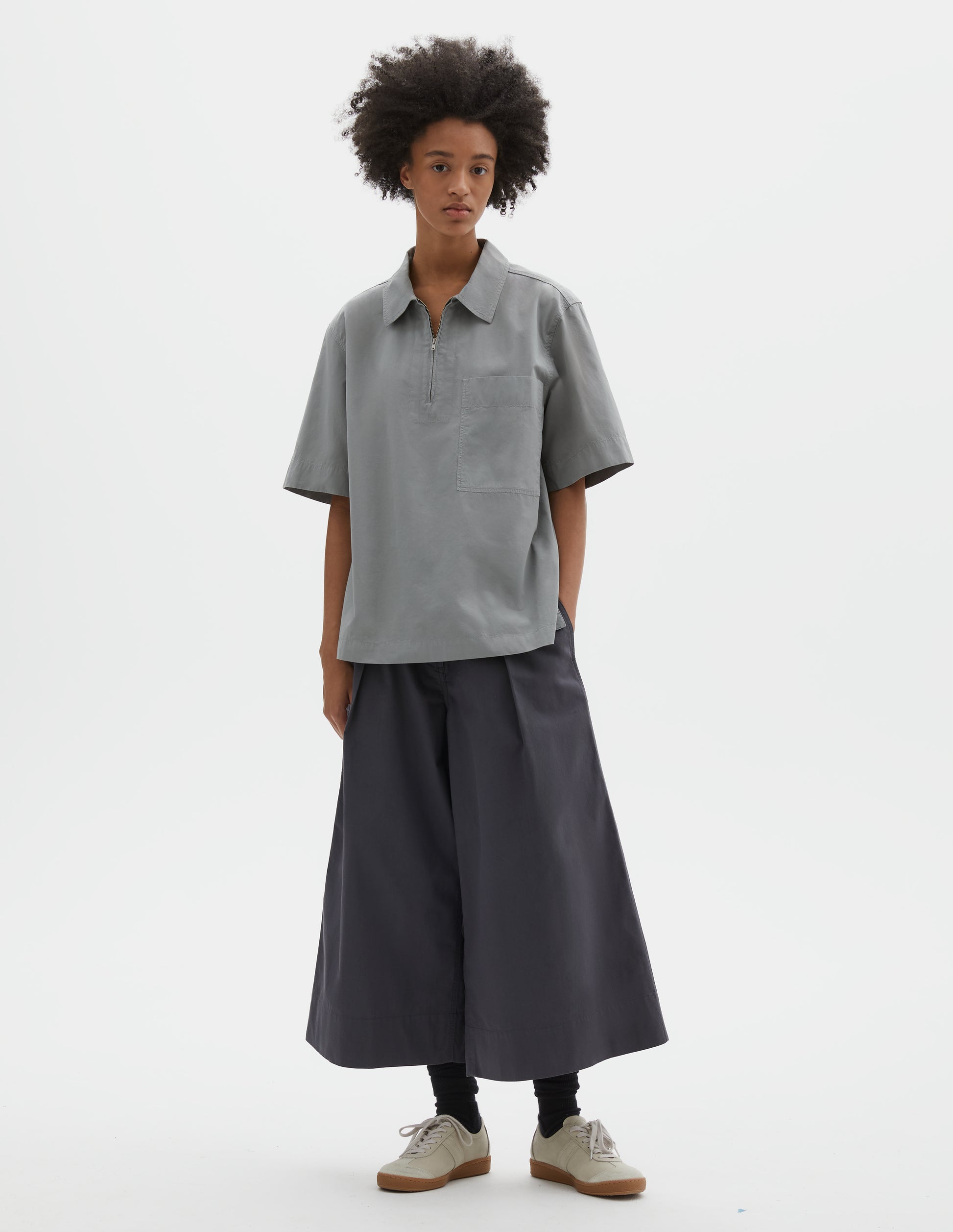 Full body image, front view of model wearing blueish grey cotton short sleeve shirt, with wide stitch through placket, half zip, patch chest pocket and side split hem. Styled with dark grey culottes, black socks and beige suede trainers.