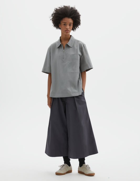 Full body image, front view of model wearing blueish grey cotton short sleeve shirt, with wide stitch through placket, half zip, patch chest pocket and side split hem. Styled with dark grey culottes, black socks and beige suede trainers.