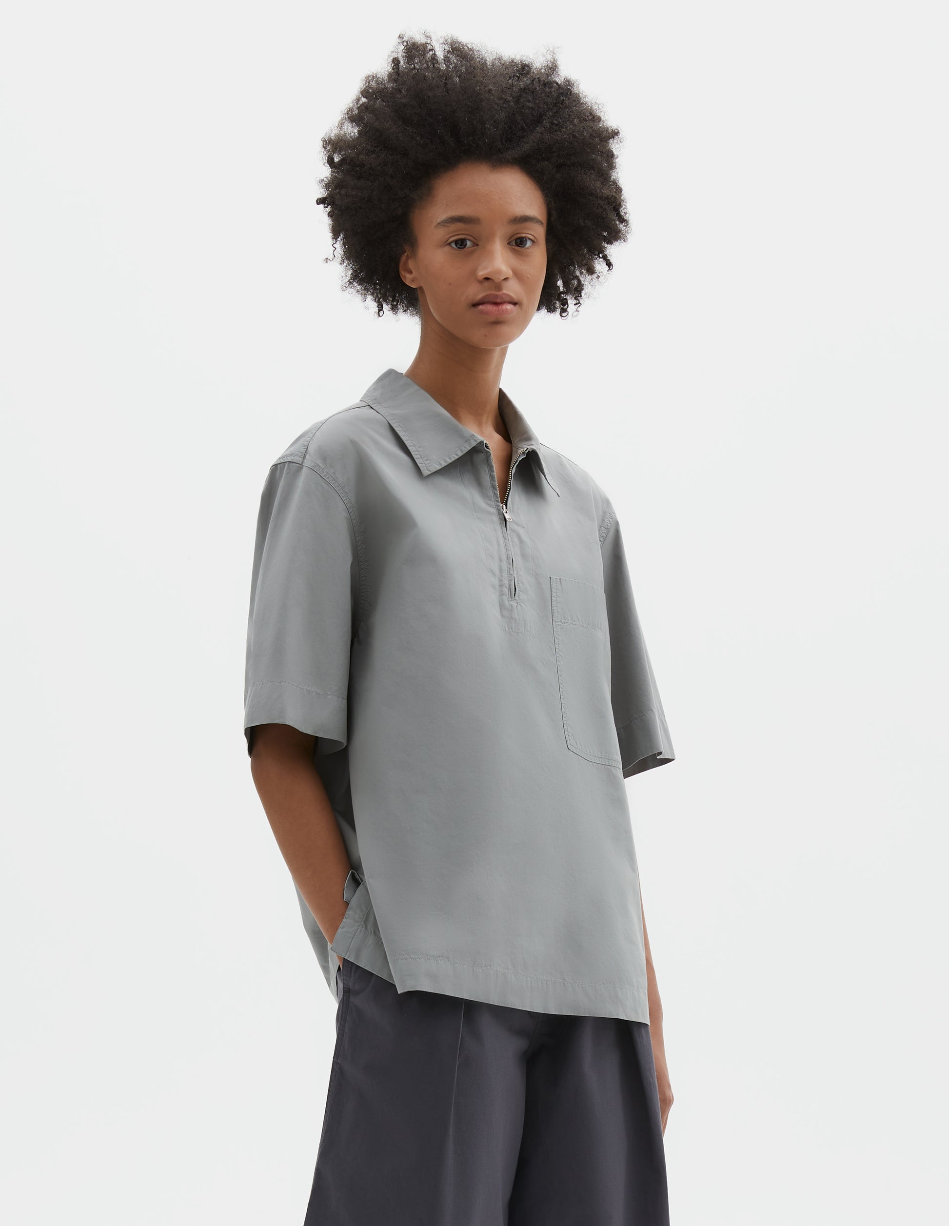 Cropped body image, side view of model wearing blueish grey cotton short sleeve shirt, with wide stitch through placket, half zip, patch chest pocket and side split hem. Styled with dark grey culottes.