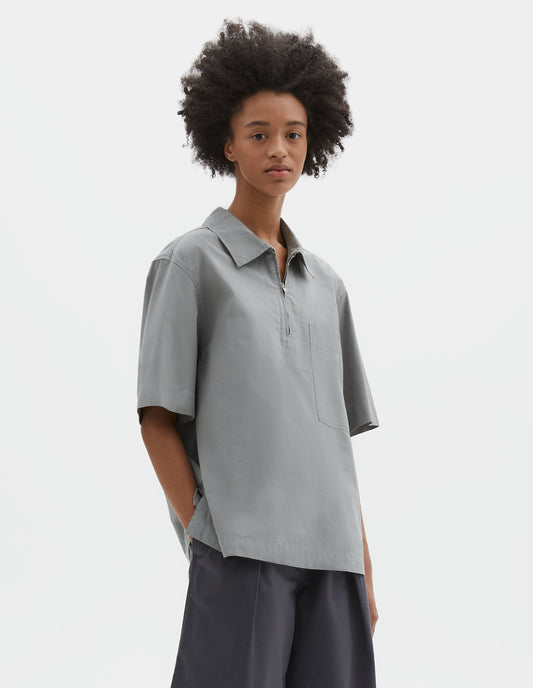 Cropped body image, side view of model wearing blueish grey cotton short sleeve shirt, with wide stitch through placket, half zip, patch chest pocket and side split hem. Styled with dark grey culottes.