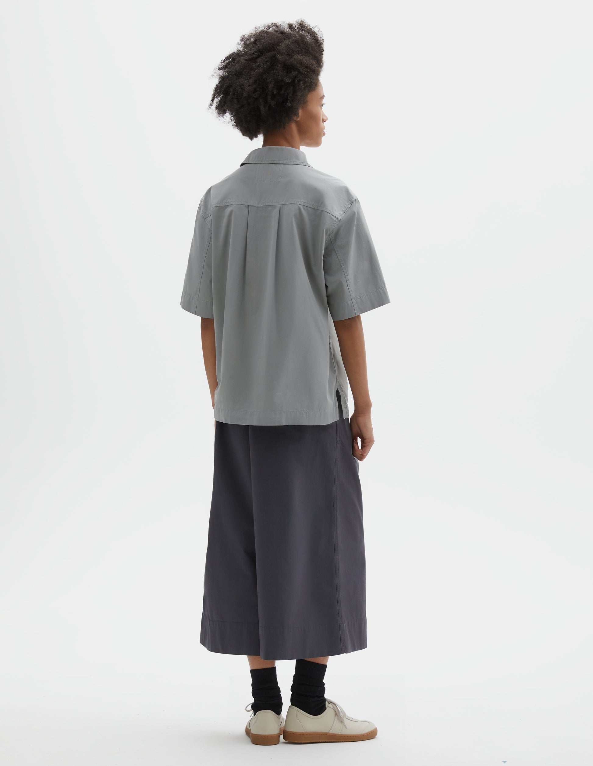 Full body image, back view of model wearing blueish grey cotton short sleeve shirt, with side split hem and wide box pleat on back yoke. Styled with dark grey culottes, black socks and beige suede trainers.