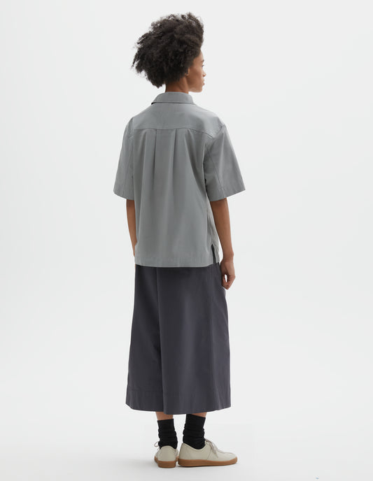 Full body image, back view of model wearing blueish grey cotton short sleeve shirt, with side split hem and wide box pleat on back yoke. Styled with dark grey culottes, black socks and beige suede trainers.