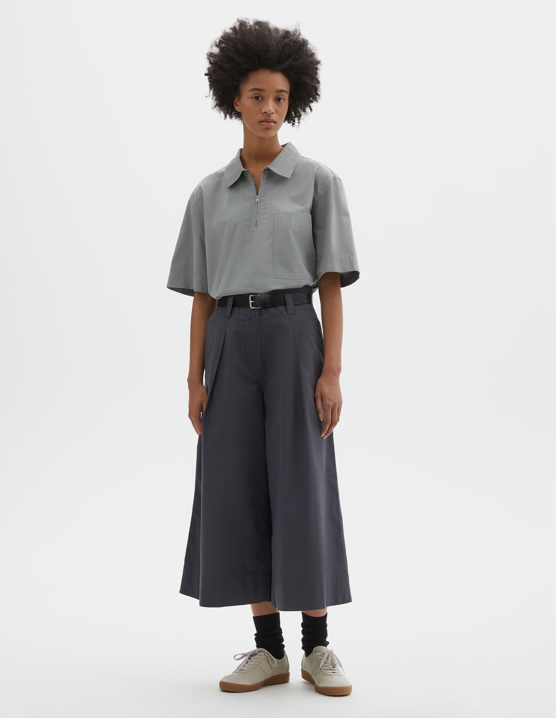 Full body image, front view of model wearing relaxed wide leg grey culottes with zip fly, stitch-through waistband with dropped belt loops, bar tacked pleats, concealed wide pockets and deep stitch-through hem. Styled with light grey top, black leather belt, black socks and beige nubuck trainers.