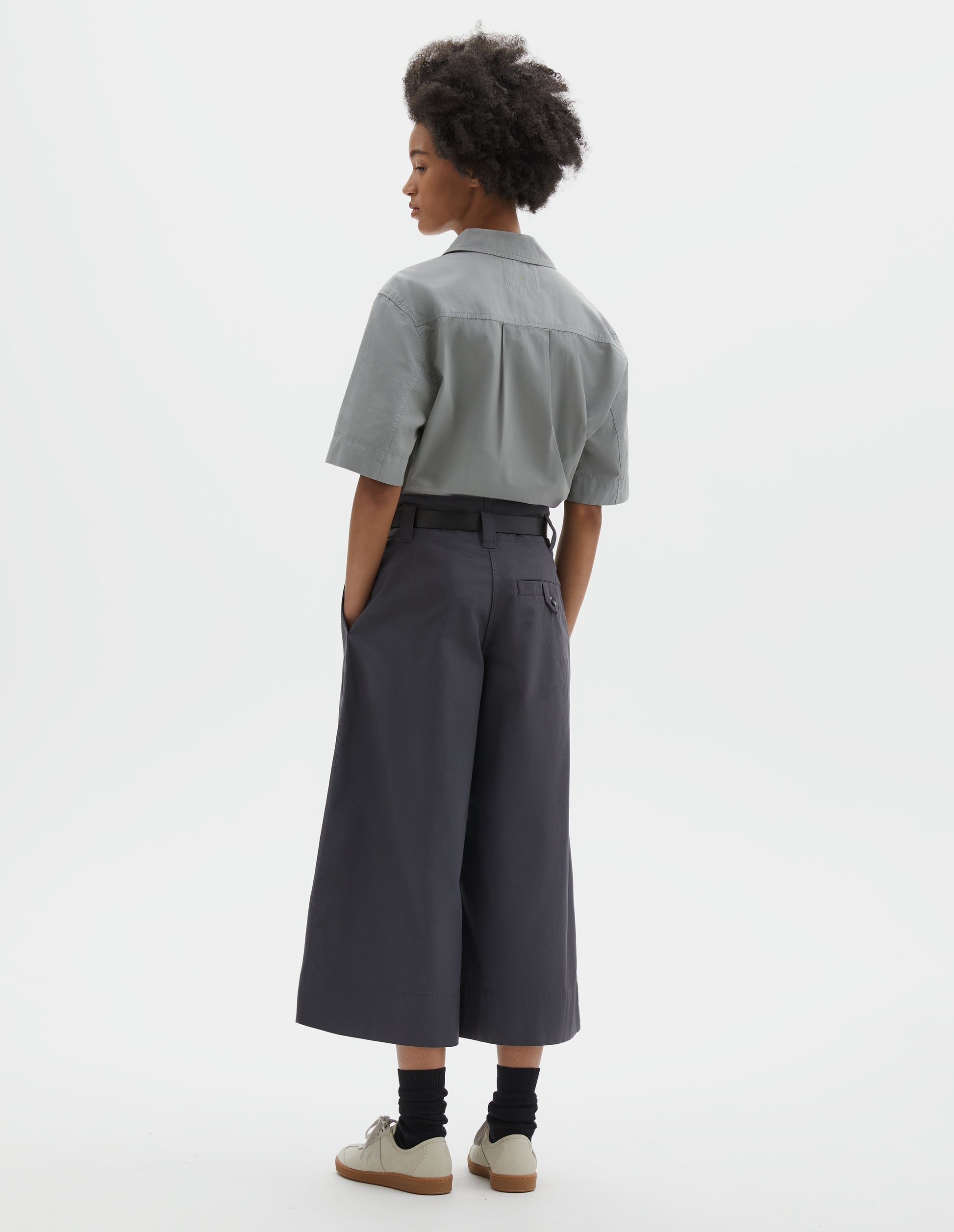 Full body image, back view of model wearing relaxed wide leg grey culottes with stitch-through waistband and dropped belt loops, and corozo button loop fastening welt pocket. Styled with light grey top, black leather belt, black socks and beige nubuck trainers.