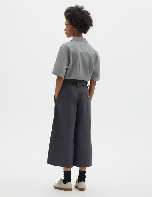 Full body image, back view of model wearing relaxed wide leg grey culottes with stitch-through waistband and dropped belt loops, and corozo button loop fastening welt pocket. Styled with light grey top, black leather belt, black socks and beige nubuck trainers.
