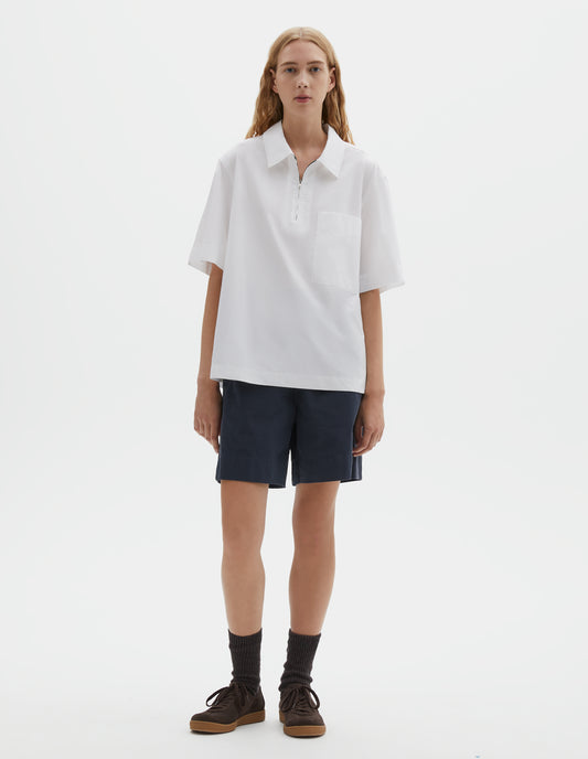 Full body image, front view of model wearing relaxed short-sleeved white shirt with wide stitch-through placket with half zip, patch chest pocket and side split hem. Styled with dark blue shorts, dark brown socks and dark brown nubuck trainers.